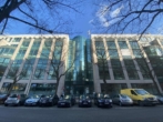 Ein modernes Bürogebäude mit Glasfront und geparkten Autos in einer städtischen Straße.