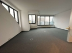 Leerer Büroraum mit grauem Teppich, großen Fenstern und Blick auf städtische Gebäude.
