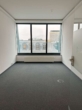 Leeres Büro mit grauem Teppich, großen Fenstern, weiß gestrichenen Wänden und Blick auf Stadtgebäude.