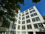 Mehrstöckiges weißes Gebäude, blauer Himmel im Hintergrund, Bäume am Rand.