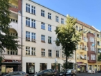 Eine vierstöckige Gebäudezeile mit Geschäften unten, farbenfrohe Fassaden, Bäume, Autos davor.