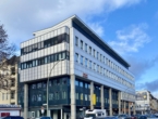 Mehrstöckiges, modernes Bürogebäude mit Glasfenstern und grauer Fassade an einer belebten Straße.
