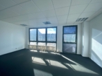 Leerer, lichtdurchfluteter Raum mit großen Fenstern und dunklem Teppichboden.