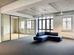 Leeres Büro mit Glaswand, blauer Ecksofa, grauer Teppichboden, große Fenster mit Blick nach draußen.