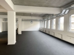 Leerer, weißer Raum mit grauem Teppichboden und großen Fenstern an der rechten Wand.