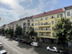 Straßenansicht mit gelbem Gebäude, Bäumen und parkenden Autos bei bewölktem Himmel.