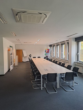 Konferenzraum mit langem Tisch, dunklen Stühlen und weißen Wänden. Fenster auf der rechten Seite.