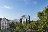 Ein modernes Gebäude mit Glasfront neben älteren Gebäuden, umgeben von grünen Bäumen.