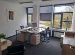 Ein leeres Büro mit weißen Wänden, grauem Teppich, Holzschreibtischen und großen Fenstern.