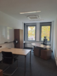 Ein leeres Büro mit Schreibtischen, Stühlen, Ventilator und Blick auf Bäume durch große Fenster.